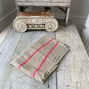 Pode incluir: Uma toalha de mesa de linho dobrada com listras vermelhas sobre uma superfície de madeira branca. A toalha de mesa está dobrada de forma que as listras vermelhas e a textura do linho são visíveis.