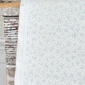 May include: Close-up of a textured, light blue and white patterned paper. The paper has a subtle, irregular pattern. The edge of the paper is visible, revealing its thickness. The paper is resting on a distressed, white-painted wooden surface.