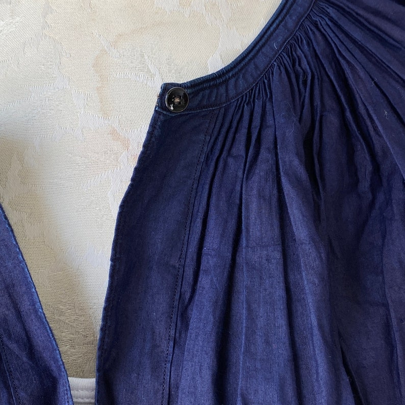 May include: Close-up of a dark blue denim shirt with a button closure at the neckline. The shirt has a gathered detail at the neckline.