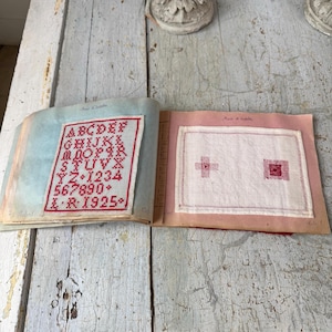 Puede incluir: Libro abierto con páginas que muestran muestras de punto de cruz rojo. Una página muestra un alfabeto y números en hilo rojo. La otra página muestra dos diseños de punto de cruz más pequeños. El libro tiene una cubierta azul claro y rosa.