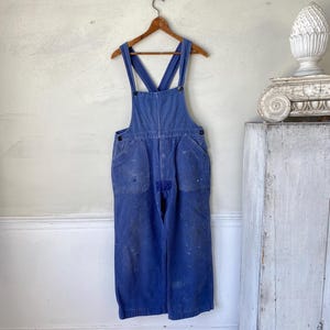 Può includere: Salopette da lavoro blu vintage appesa a una gruccia di legno. La salopette ha un davanti a pettorina, tasche e spalline regolabili. Il tessuto mostra segni di usura e macchie di vernice, conferendole un aspetto rustico e vissuto. È uno stile classico di abbigliamento da lavoro.
