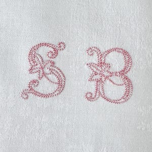 May include: White fabric with pink embroidered letters "S" and "B" in a cursive script.