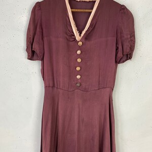 May include: A long, burgundy silk dress with a pink satin trim around the neckline. The dress has a V-neckline and short sleeves. It features a row of buttons down the front.