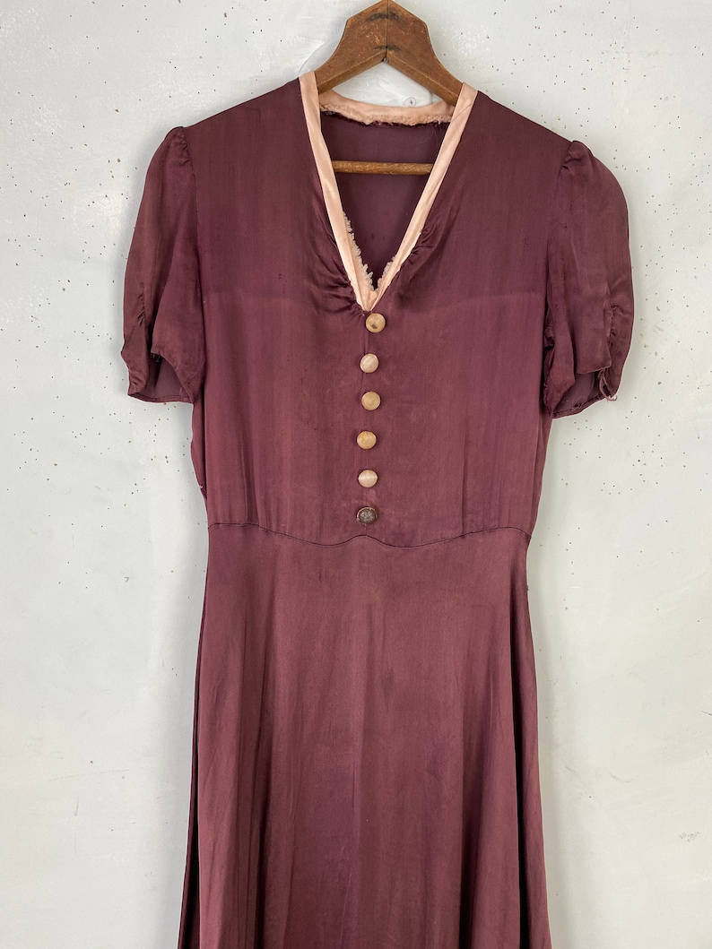May include: A long, burgundy silk dress with a pink satin trim around the neckline. The dress has a V-neckline and short sleeves. It features a row of buttons down the front.