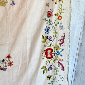 May include: A white linen tablecloth with a hand-stitched embroidered border featuring a floral design in shades of red, green, blue, and purple. The embroidery includes flowers, leaves, and butterflies.