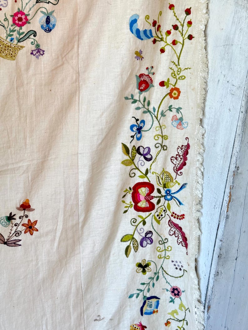 May include: A white linen tablecloth with a hand-stitched embroidered border featuring a floral design in shades of red, green, blue, and purple. The embroidery includes flowers, leaves, and butterflies.