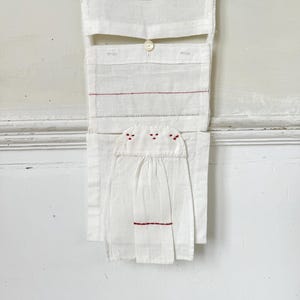 May include: White fabric wall hanging with red stitching and a button. The fabric is folded and layered to create pockets and a unique design.