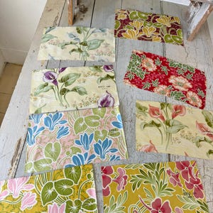 May include: Assortment of eight fabric swatches featuring floral patterns in various colors, including yellow, green, pink, red, and blue. The fabric swatches are arranged on a wooden surface.