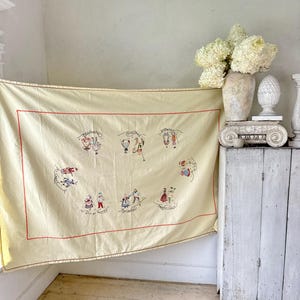 May include: A cream-colored tablecloth with red stitching and embroidered images of people in traditional clothing. The tablecloth features the months of the year in French.