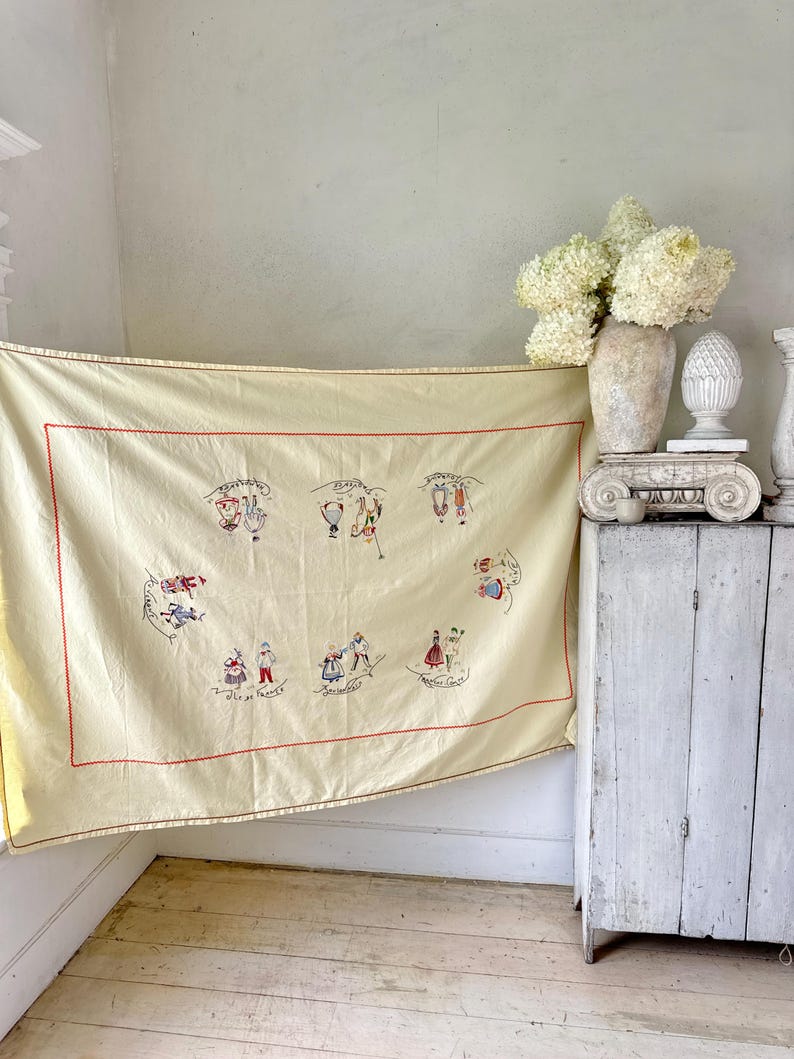 May include: A cream-colored tablecloth with red stitching and embroidered images of people in traditional clothing. The tablecloth features the months of the year in French.