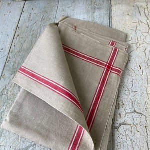 Pode incluir: Um conjunto de duas toalhas de chá de linho com listras vermelhas. As toalhas estão dobradas e empilhadas sobre uma superfície de madeira branca.