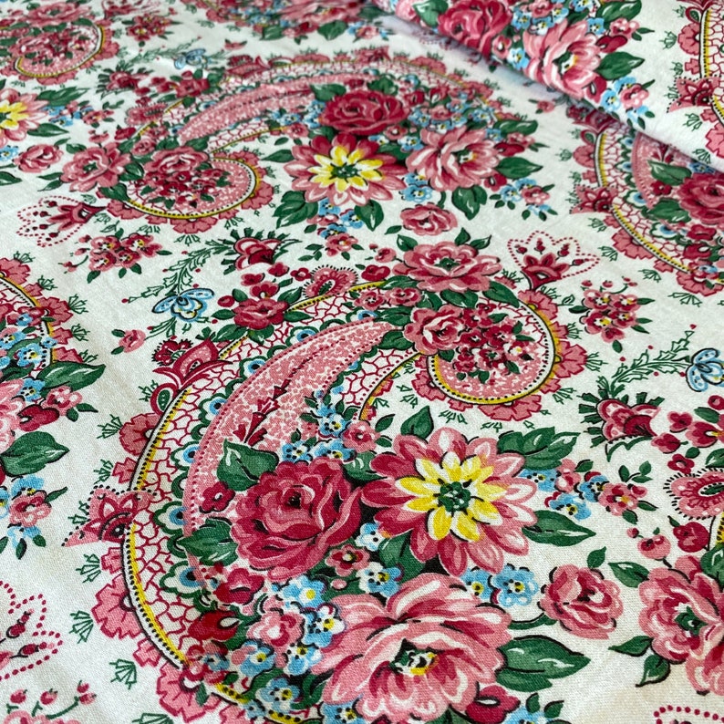May include: Close-up of a floral fabric with a paisley design. The fabric features a white background with a repeating pattern of pink roses, yellow and blue flowers, and green leaves. The paisley elements are in pink and red.