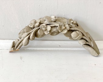 May include: A gray, decorative, floral-shaped molding with a curved design. The molding is made of plaster or a similar material and features a series of stylized flowers and leaves.