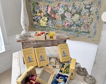 May include: A still-life arrangement featuring vintage embroidery supplies. Boxes of colorful embroidery floss and thread are displayed alongside vintage books. A floral tapestry hangs in the background, adding a touch of elegance to the scene.