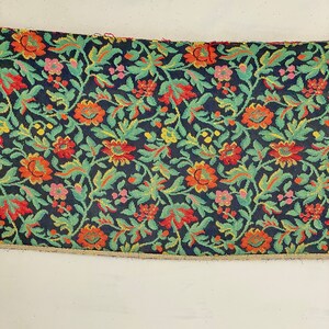 May include: A close-up of a fabric with a floral pattern. The fabric is dark blue with green, red, orange, and yellow flowers. The fabric has a textured surface.