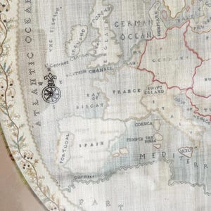 以下が含まれることがあります： フランス、スペイン、ポルトガルなどの国々を含むヨーロッパの詳細な地図が、テクスチャ加工されたオフホワイトの生地に表示されています。 地図には地理的特徴とテキストラベルが含まれており、隅には羅針盤があります。