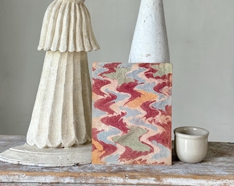 Può includere: Un blocco rettangolare con un motivo ondulato colorato nei toni del rosso, blu e giallo. Accanto, una piccola tazza in ceramica bianco sporco, un'alta colonna decorativa bianca e una statua scolpita color bianco sporco.