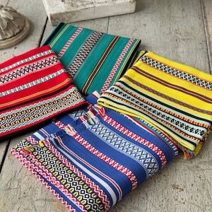 Puede incluir: Textiles plegados en rojo, verde, amarillo y azul con intrincados patrones de rayas y geométricos. Los textiles tienen una textura tejida y están dispuestos sobre una superficie de madera envejecida. Los diseños incorporan varios colores y formas.