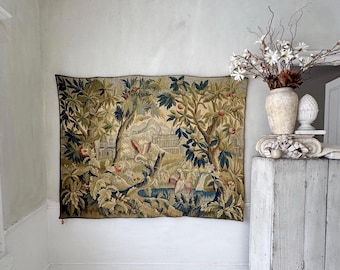 May include: A large tapestry depicting a forest scene with trees, a body of water, and architectural elements. The tapestry features a color palette of greens, browns, and blues. It is hanging on a white wall in a room with a wooden floor.