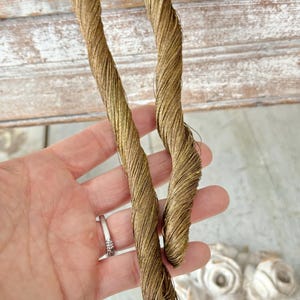 May include: Close-up of two strands of gold-colored twisted thread. The thread is held in a hand, with a ring visible on a finger. The thread appears to be made of a fine, shiny material, possibly silk or metallic thread. The background is a blurred view of a white decorative element.