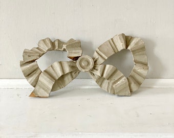 Puede incluir: Un lazo de madera decorativo con un diseño fruncido, pintado en gris claro.
