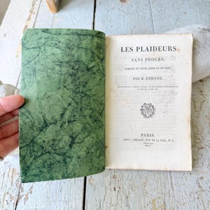 May include: An open antique book titled Les Plaideurs Sans Procès by M. Étienne, published in Paris in 1821. The left page features a textured green marbled endpaper, while the right page displays black printed text and a small circular emblem.