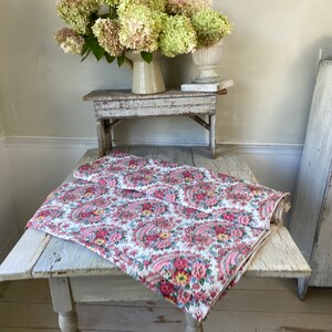 May include: A folded floral paisley patterned fabric in shades of pink, red, and yellow on a white background. The fabric is displayed on a distressed white wooden table. A vase of flowers sits in the background.