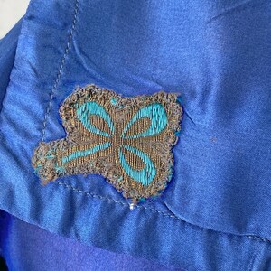 May include: Close-up of a blue satin fabric with a decorative butterfly-shaped appliqu&eacute;. The appliqu&eacute; features a gray background with teal and gold embroidery, adding a touch of elegance to the fabric.