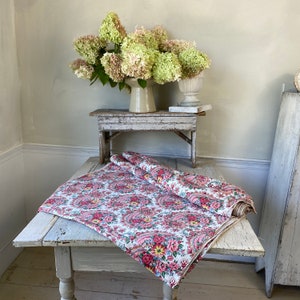 May include: A white tablecloth with a pink paisley floral pattern is draped over a distressed, white wooden table. A rolled-up section of the fabric sits on top. A bouquet of green and pink flowers sits in a vase on a small wooden table.