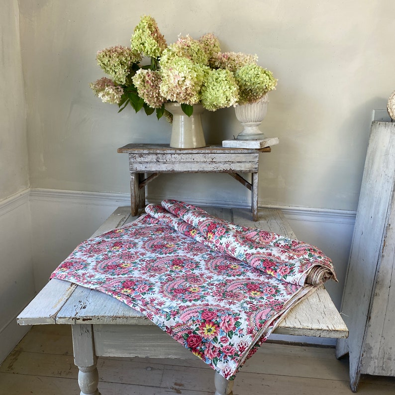 May include: A white tablecloth with a pink paisley floral pattern is draped over a distressed, white wooden table. A rolled-up section of the fabric sits on top. A bouquet of green and pink flowers sits in a vase on a small wooden table.