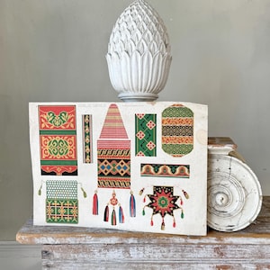 May include: A vintage printed sheet with colorful geometric patterns and designs. The sheet features a variety of borders, tassels, and decorative elements. The sheet is displayed on a wooden table with a white ceramic pineapple finial.