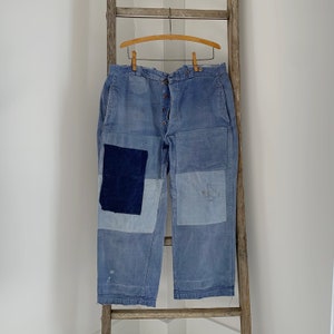 Może przedstawiać: Spodnie z niebieskiego denimu z kilkoma naszytymi łatkami. Spodnie mają zapięcie na guziki i wiszą na drewnianym wieszaku.