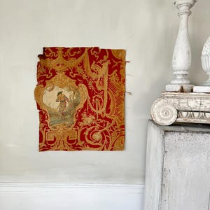 May include: A vintage red and gold fabric with a floral pattern and a central image of a person in a boat. The fabric is torn and faded, suggesting it is antique.