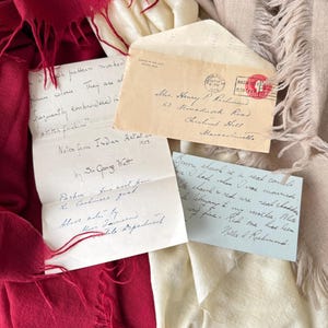 Puede incluir: Una vista aérea presenta textiles y correspondencia manuscrita. Se observa una bufanda con flecos de color rojo intenso, junto con telas crema y beige. Las cartas incluyen un sobre con matasellos y notas manuscritas.