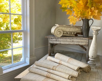 Puede incluir: Una mesa de madera rústica con un acabado blanqueado, que presenta un elemento arquitectónico decorativo de yeso blanco y una pila de tela de lino natural con rayas marrones. La mesa está colocada contra una pared blanca con una ventana detrás.