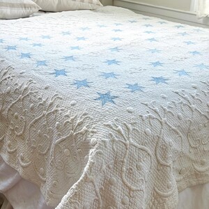 Peut inclure: Une courtepointe blanche ornée d'étoiles bleues et de broderies florales détaillées. La courtepointe est posée sur un lit avec des oreillers blancs et un jupon de lit blanc. La texture de la courtepointe est visible, mettant en valeur les coutures.