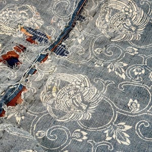 May include: A close-up of a blue and white fabric with a repeating pattern of stylized birds and floral designs. The fabric has a distressed look, with some areas showing wear and tear.