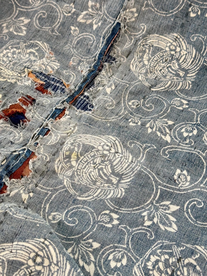 May include: A close-up of a blue and white fabric with a repeating pattern of stylized birds and floral designs. The fabric has a distressed look, with some areas showing wear and tear.