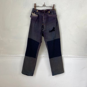以下が含まれることがあります： 膝に黒い布のパッチが付いた濃いグレーの作業ズボン。ボタンフライで、使い古して色あせています。