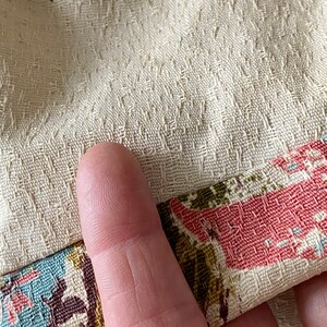 May include: Close-up of a cream-colored fabric with a textured pattern. The fabric has a pink and green floral print on the bottom edge.
