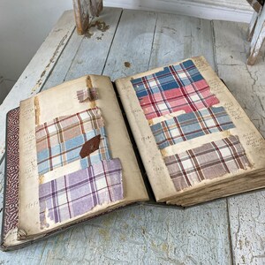 May include: An open book with pages filled with different plaid fabric swatches. The swatches are in various colors, including red, blue, and brown. The book is open to reveal the swatches and has a worn, vintage look.