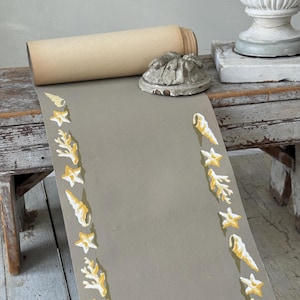 May include: A rolled-up, tan paper scroll rests on a weathered wooden surface, next to a gray decorative object. A gray runner is decorated with yellow and white seashell, starfish, and coral designs. The runner is unrolled, revealing its decorative border.