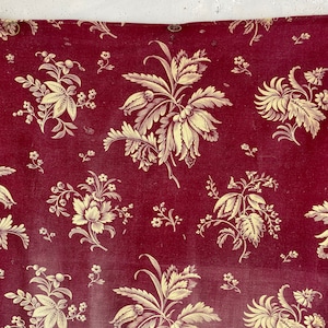 Könnte beinhalten: Ein burgunderfarbener Stoff mit einem sich wiederholenden Muster aus goldenen Blumendesigns. Der Stoff hat eine leicht strukturierte Oberfläche.