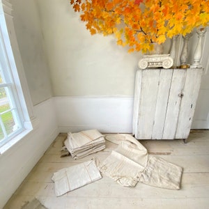 May include: A stack of white linen fabric with brown stripes, folded and arranged on a light wood floor. The fabric is in a room with white walls and a white cabinet with a decorative piece on top.