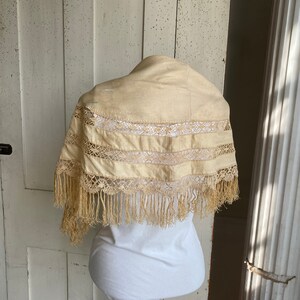May include: A cream-colored lace shawl with fringe trim. The shawl is displayed on a white mannequin.