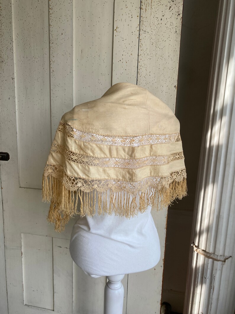 May include: A cream-colored lace shawl with fringe trim. The shawl is displayed on a white mannequin.