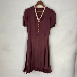 May include: A burgundy silk dress with a pink lace trim around the neckline and short sleeves. The dress has a fitted bodice and a flowing skirt with a ruffled hem. The dress has five brown buttons down the front.