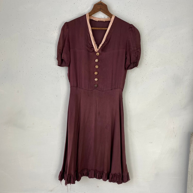 May include: A burgundy silk dress with a pink lace trim around the neckline and short sleeves. The dress has a fitted bodice and a flowing skirt with a ruffled hem. The dress has five brown buttons down the front.