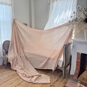 May include: A peach-colored, striped bedspread draped over a vintage bed frame. The fabric has a subtle vertical stripe pattern. The scene is set in a room with wooden floors, a fireplace, and a decorative chair, suggesting a vintage or farmhouse aesthetic.