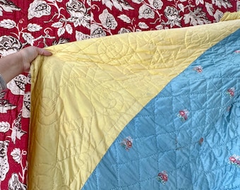 May include: A quilted blanket with a yellow and blue design. The yellow section has a quilted pattern, while the blue section features small floral embroidery. The background is a red floral pattern.